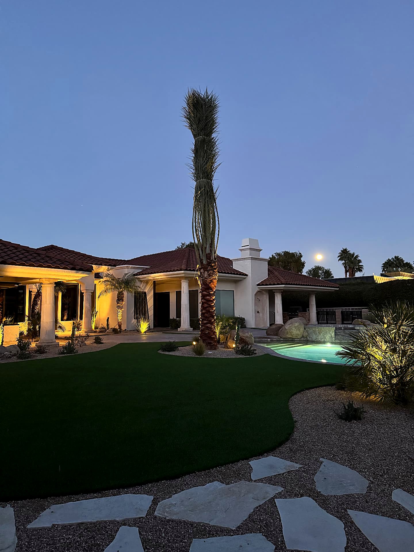 Twilight backyard with uplit palms, lit pool and moonrise over the roofline
